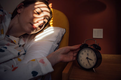Woman in heart-print pajamas waking in bed, reaching for a black alarm clock on a bedside table as morning sunlight from blinds falls across her face