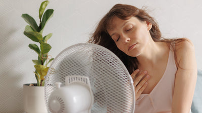 Woman cooling off in front of a tabletop fan at home, eyes closed and hand on chest, potted plant in background