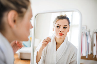 Young woman in a white robe looking at her reflection in a bedroom mirror while adjusting her hair in soft morning light